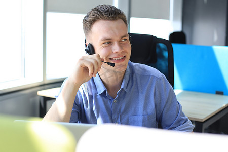 性格开朗的年轻人在工作场所使用耳机支持电话男接线员，同时使用笔记本电脑、帮助服务和客户咨询呼叫中心概念。