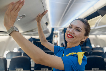 身穿蓝色制服的漂亮空姐的画像，在关闭手提行李舱时对着镜头微笑，留在飞机内的过道上