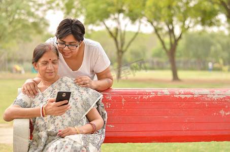 印度新德里的一个公园里，年轻的印度女孩把手放在印度孟加拉老母亲的肩膀上，坐在红色长凳上看着手机，忙着说话。