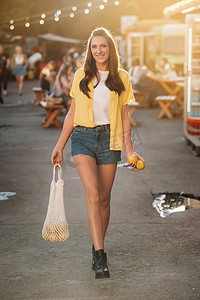 喝果汁的女孩摄影照片_一个身穿黄色衬衫、短裤和黑色鞋子的女孩，带着一网柠檬和柠檬汁，正在城市街道上行走。
