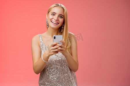 迷人优雅的 25 多岁年轻女性穿着银色连衣裙，在夜总会朋友聚会后，拿着智能手机消息，使用应用程序订购出租车，开心地站着，无忧无虑地微笑着拍照