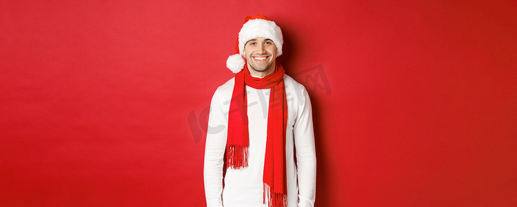 庆祝圣诞节和微笑的快乐白种人，戴着圣诞帽、围巾和白色毛衣，享受新年假期，站在红色背景上