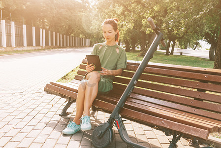 数字生态摄影照片_女孩在公园里用数字平板电脑和电动滑板车坐在长凳上休息。