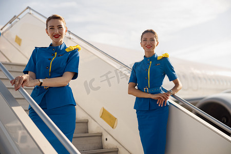 两位身穿蓝色制服的漂亮空姐对着镜头微笑，站在楼梯上欢迎乘客