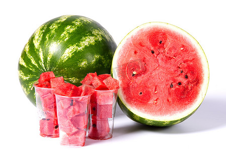 红色的立方体摄影照片_在新鲜有机绿色西瓜前面的塑料杯中切成红色西瓜块，并切成一半具有红色质地的西瓜。
