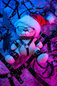 游戏皮肤摄影照片_一只戴着圣诞老人帽子的熊是 BDSM 游戏的圣诞礼物，来自一家霓虹灯下的性用品商店，供新年使用