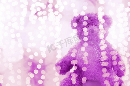 泰迪熊娃娃在灯光线散景紫色明亮的圣诞节或新年快乐背景，熊孤独地坐在闪闪发光的紫色白色背景模糊的散景明亮（选择性焦点）