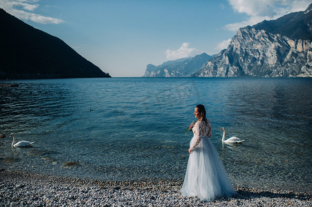 加衣服摄影照片_一个穿着漂亮的白色连衣裙的女孩沿着加尔达湖的堤岸散步。一个女人在意大利的一座山和湖的背景下被拍照。Torbole