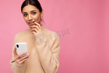 特写照片中，穿着米色温暖毛衣的漂亮可爱的黑发年轻女子站在粉色背景上，通过电话在互联网上冲浪，看着相机，感到担忧