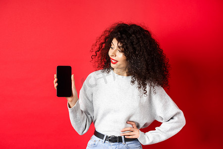 一头卷发的时尚年轻女性的肖像显示空的智能手机屏幕，展示购物应用程序，站在红色背景上
