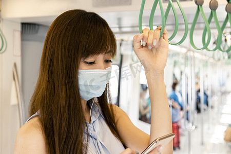 亚洲年轻女乘客戴着外科口罩，在地铁里用手机听音乐