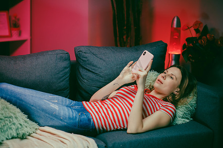 轻松的女孩拿着智能手机使用移动应用程序观看有趣的视频笑着躺在沙发上，微笑着懒惰的年轻女人在家里的沙发上在社交媒体上聊天很开心