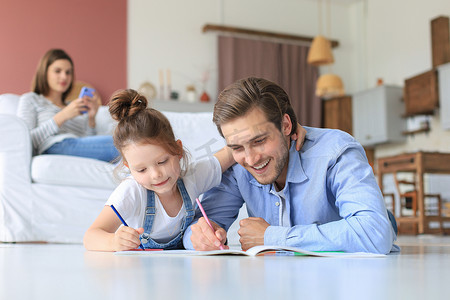 快乐的父亲微笑着女儿躺在温暖的地板上享受创造性活动，在相册中画铅笔着色图片，母亲在沙发上休息，家人一起度过空闲时间。