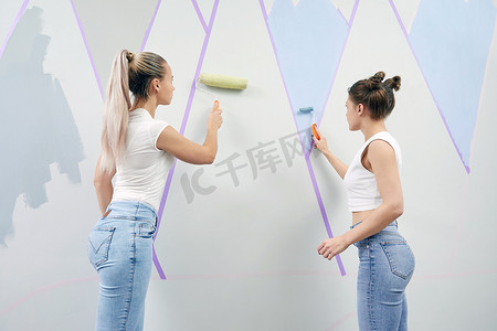 两名年轻女性用油漆滚筒和遮蔽胶带粉刷墙壁