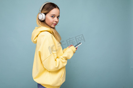 身穿黄色时尚连帽衫、蓝色背景中突显的侧脸漂亮年轻金发女性，戴着白色蓝牙无线耳机，听着酷炫的音乐，用手机上网看着相机