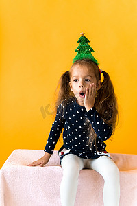 鹿黄色摄影照片_正面微笑的肖像惊讶快乐的小女学生女孩圣诞树装饰在头上的圆点连衣裙上竖起大拇指橙色背景。