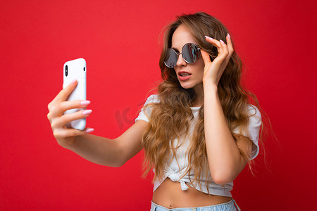 令人惊叹的美丽年轻女子拿着手机自拍照片使用智能手机相机戴着太阳镜日常时尚服装隔离在彩色墙壁背景上看着设备屏幕