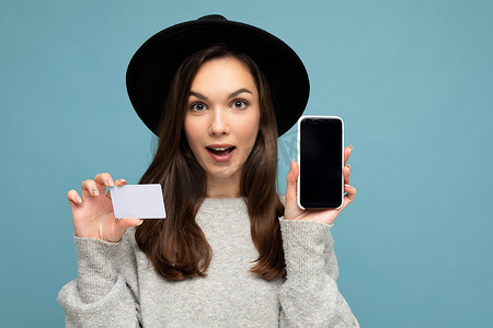 身穿黑色帽子和灰色毛衣的美丽黑发年轻女性，与蓝色背景隔离，手持信用卡和手机，显示屏空空如也，用于模拟看相机