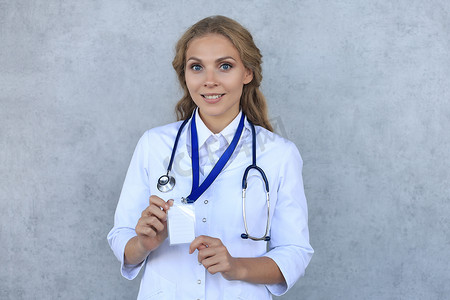 专业徽章摄影照片_微笑的金发女医生穿着制服站在灰色背景中，在徽章上显示她的名字。