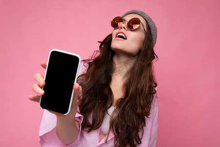 正面迷人的黑发年轻女性，戴着时尚的粉色衬衫灰色帽子，戴着彩色太阳镜，在粉色背景中显得格格不入，手里拿着手机，展示着空显示屏的手机，供模型查找