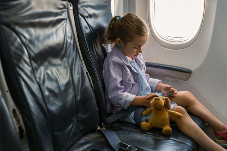 坐飞机的孩子摄影照片_乘飞机旅行时坐在椅子上拿着玩具拿着电话的小女孩