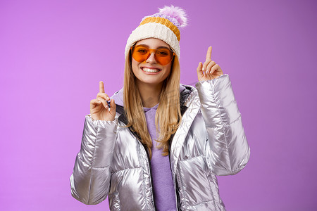 快乐、精力充沛、娱乐的可爱金发女人，享受假期雪山之旅，戴着墨镜，银色夹克，冬帽，指着逗乐的站立紫色背景跳舞