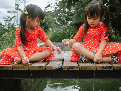 运动角色儿童摄影照片_池塘边的木桥上，两个穿着红裙子的小妹扮演着钓鱼的角色。
