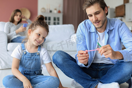 快乐的父亲微笑着女儿躺在温暖的地板上享受创造性活动，在相册中画铅笔着色图片，母亲在沙发上休息，家人一起度过空闲时间。