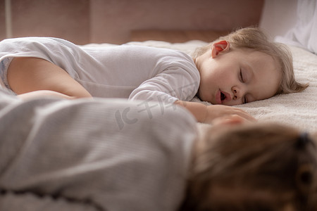 童年、睡眠、放松、家庭、生活方式概念 — 两个 2 岁和 3 岁的小孩穿着白色和米色紧身衣裤，午餐时睡在米色和白色床上，手牵着手俯视。