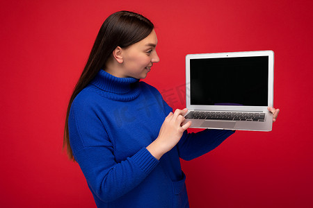 红墙背景中，身穿蓝色毛衣的漂亮年轻黑发女性拿着笔记本电脑看着上网本键盘的照片