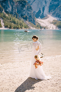 美丽的新娘穿着白色的袖子和蕾丝连衣裙，带着一束黄色的秋干花和牡丹玫瑰，在意大利的布拉伊斯湖上。