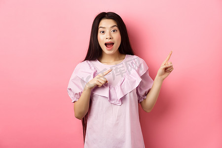 兴奋的亚洲少女指着右手，笑得开心又开心，展示酷新闻，站在粉红色背景上