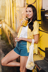 一个身穿黄色衬衫、短裤和黑色鞋子的女孩，带着一网柠檬和柠檬汁，正在城市街道上行走。