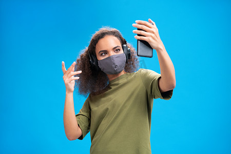 在视频通话中，年轻的非洲裔美国女孩使用她的智能手机，戴着耳机，穿着橄榄色 T 恤、可重复使用的面罩，以防止其他人感染蓝色背景中突显的 Corona COVID-19