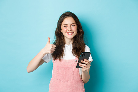 美丽快乐女孩的肖像，竖起大拇指，手持智能手机，网上购物，在网上下订单，看起来很满意，称赞好应用，蓝色背景