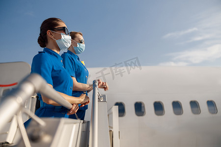 两名身穿蓝色制服、戴着墨镜和防护面罩的年轻空姐在登机时站在楼梯上，望向别处的侧视图