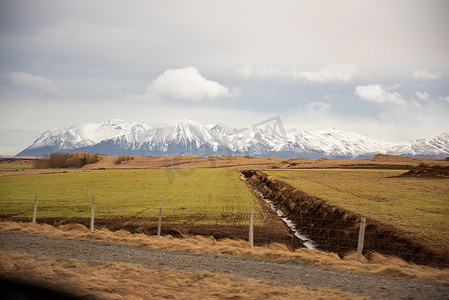 山海的线条摄影照片_美丽的绿色和橙色的冰岛地形与水路形成的线条通向一个蓝色的白雪皑皑的山脉，蓬松的云彩引人注目的天堂般的农田围栏。