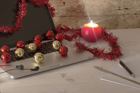 圣诞假期的电脑工作概念：打开的铝制笔记本电脑、红色花环装饰、红色和金色的小玩意、点燃的蜡烛、钢笔和尺子，放在浅色木桌上的方块笔记上
