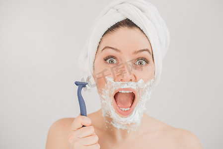 脸上的泡沫摄影照片_一个女人脸上涂着剃须泡沫的滑稽肖像，她拿着一把剃须刀，背景是白色的。