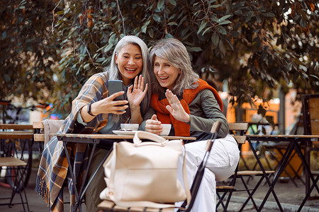 积极的成熟女性朋友在街头咖啡馆通过智能手机在视频聊天中挥手