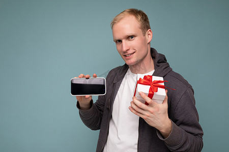 一张帅气、面带微笑的年轻金发男子被隔离在蓝色背景墙上，穿着休闲时尚的衣服，手拿带红丝带的白色礼盒和带空屏幕显示的手机，用于模拟看相机