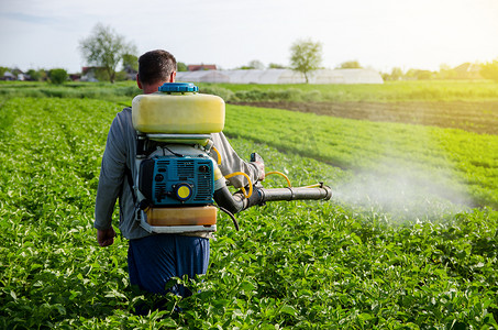 农业喷雾器摄影照片_一位农民用雾化喷雾器在马铃薯灌木丛上喷洒杀菌剂和杀虫剂。