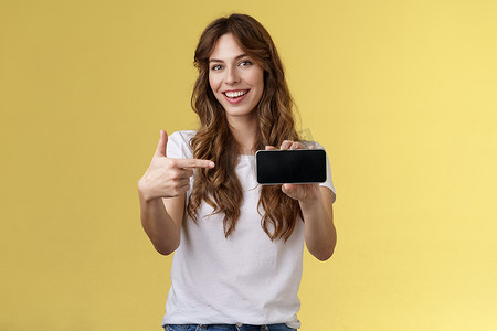 自信时髦的漂亮时尚女性介绍很棒的游戏应用程序展示智能手机指向食指空白手机屏幕微笑高兴推荐尝试应用程序黄色背景