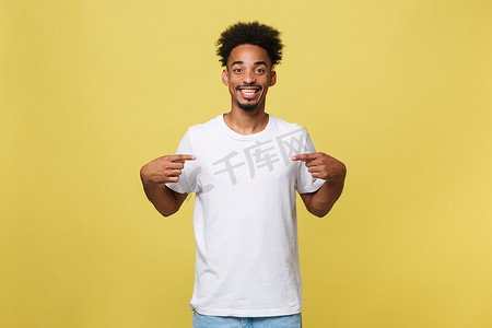 手势指示摄影照片_帅气开朗的深色皮肤男性卖衣服的画像，穿着休闲的白色 T 恤，在空白的复制空间指示您的标志，隔离在黄色背景中。