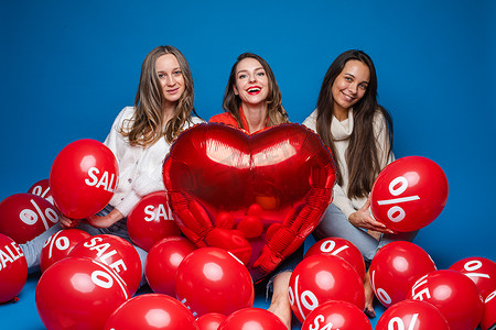快乐的女朋友摆着红色心形气球和蓝色背景上带有百分比和销售字母的气球