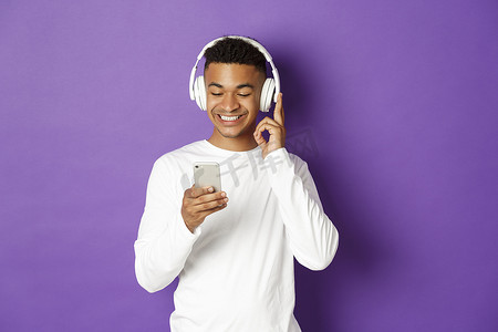 紫色背景音乐摄影照片_身穿白色运动衫、戴着耳机听音乐、看着手机、站在紫色背景上的现代非洲裔美国人形象