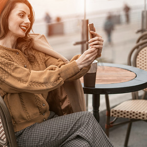 美丽的年轻女子坐在春天的户外咖啡馆里喝着咖啡，用智能手机进行视频通话。
