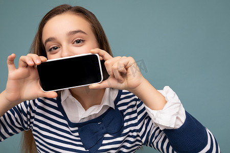 身穿条纹长袖的相当积极的黑发女孩的特写照片，站在蓝色背景上，与世隔绝，复制空间拿着智能手机，手里拿着手机，屏幕显示空，用于模型指向 gadjet 看着相机
