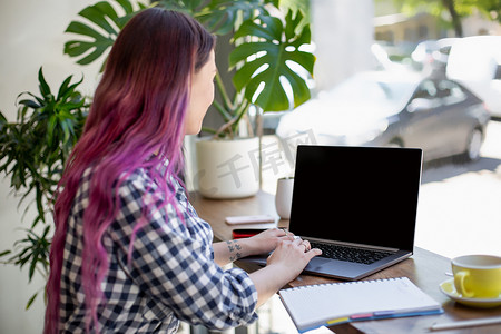 一个年轻的粉红色头发女人坐在咖啡馆里用空白的复制空间屏幕在笔记本电脑上敲键盘的背影。