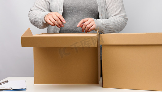 打开的盒子盒子摄影照片_穿着灰色衣服的女人站着，拿着白色背景上打开的棕色盒子，移动、发送和交付货物